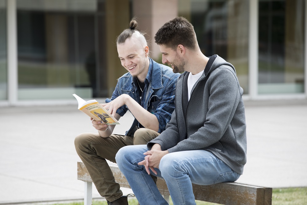 Grafiikassa kaksi miestä lukee kirjaa istuen penkin selkänojalla.