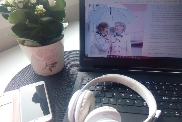 Kuvassa kannettavan tietokone, jonka näppäimistön päällä kuulokkeet. Vierellä kukka ja puhelin.